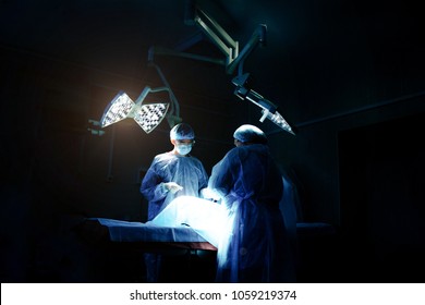 Surgery In A Clinic By A Group Of Surgeons Under A Lamp In A Dark Room