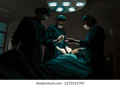 Surgical team performing surgery to patient in sterile operating room. In a surgery room lit by a lamp, a professional and confident surgical team provides medical care to an unconscious patient. - Powered by Shutterstock