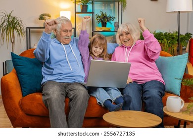 Surprise Caucasian grandfather, grandmother and granddaughter using laptop netbook while celebrating success at home. Happy girl with grandparents amazed by online game win clenching fists on sofa. - Powered by Shutterstock