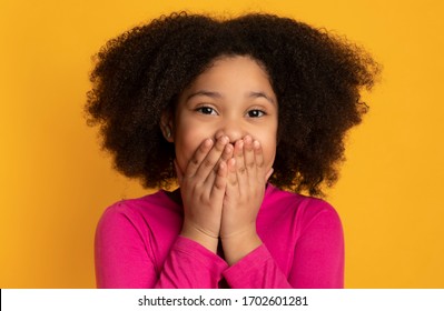 Surprise Emotion. Cute little black girl covering mouth with hands, posing over yellow background in studio, free space