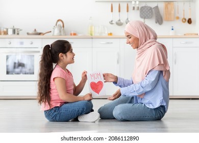 Surprise For Mom. Adorable Little Girl Giving Handmade Greeting Card To Muslim Mommy At Home, Congratulating With Mother's Day While They Sitting Together On Floor In Kitchen, Side View Shot