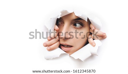 Surprise. Portrait of a young caucasian woman looking through the hole in white paper. Distrustful look. Women's curiosity and gossip. A jealous wife. Copy space.