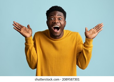 Surprised african american stylish young guy in yellow looking at camera with open mouth and raising his palms up, gesturing over blue studio background, showing amazement, closeup shot