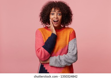 Surprised African American woman with an afro hairstyle with amazement looks camera, palm holds her cheek, feels impressed, looks affected excited overwrought, isolated on a pink background