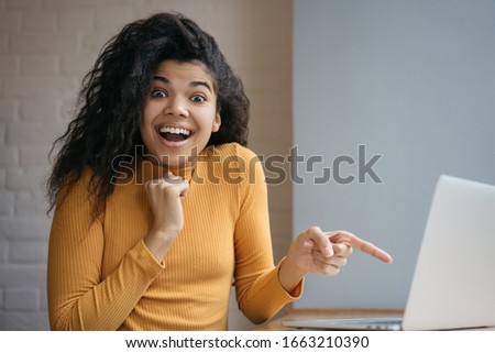 Surprised African American woman showing finger on laptop screen. Portrait of excited successful blogger influencer looking at camera with open mouth and happy emotional face  