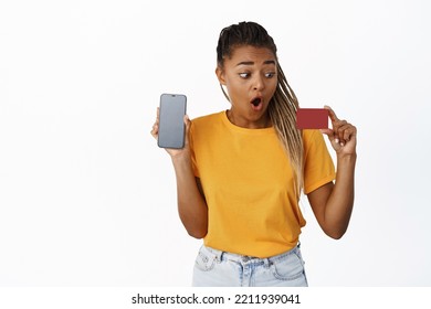 Surprised African Girl Looking At Credit Card Amazed, Showing Smartphone Screen, Concept Of Money And Finance, Technology, Standing Over White Background