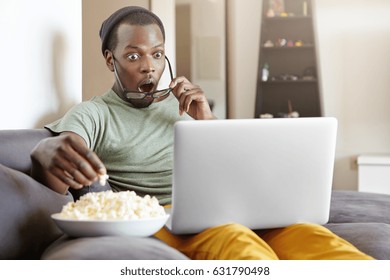 Surprised African male sitting on couch at home, eating popcorn and watching exciting TV show online on laptop computer or shocked with cliffhanger ending of detective series, keeping his mouth open