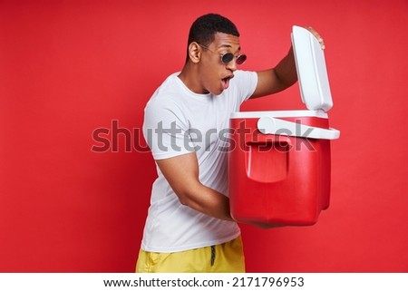 Surprised African man looking inside of cooler box while standing against red background