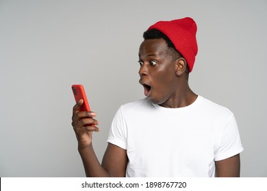 Surprised Afro Young Man Makes Big Eyes Opens Mouth Widely, Shocked With Unprecedentedly Low Prices, Indecent Photo Or Message, Looking At Mobile Phone, Isolated On Studio Grey Background. Amazed Face