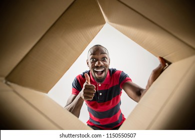 The Surprised Afrocan Man Unpacking, Opening Carton Box And Looking Inside. The Package, Delivery, Surprise, Gift Lifestyle Concept. Human Emotions And Facial Expressions Concepts