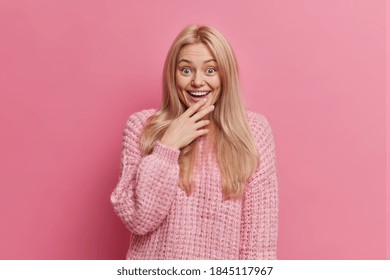 Surprised amused blonde woman stares at something wonderful with broad smile looses speech from amazement dressed in warm winter sweater isolated over rosy background cannot believe in her achievement