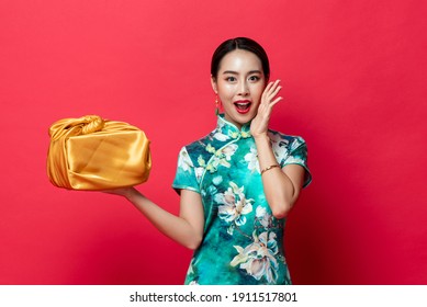 Surprised Asian woman in green traditional Cheongsam dress holding gold gift box in isolated studio red background for Lunar new year concepts