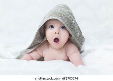 Surprised Baby Girl Or Boy After Shower With Towel On Head In White Sunny Bedroom. Child With Big Blue Eyes Open Mouth And Crying In Bed After Bath Or Shower