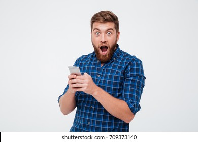 Surprised bearded man in checkered shirt using smartphone and looking at the camera over gray background