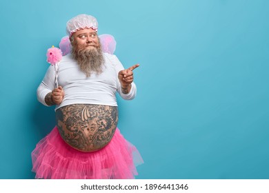 Surprised Bearded Man With Fat Tattoed Belly Wears Fairy Costume Holds Magic Wand Indicates Away With Interest Shows Something For Your Party Or Holiday Isolated On Blue Background For Your Promotion