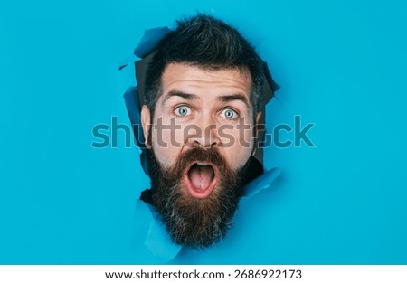 Surprised bearded man looking through paper hole. Handsome guy with stylish hairstyle making hole in paper wall. Male head looking through hole. Seasonal sale and discount. Copy space for advertising