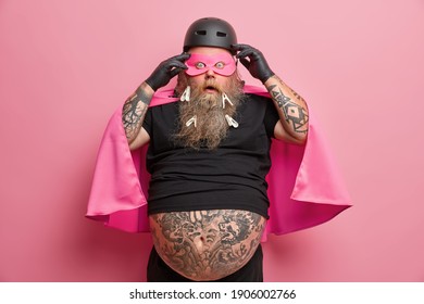 Surprised Bearded Man Looks Embarrassed Wears Superhero Costume Stares At Camera Prepares For Organizing Childrens Holiday Or Costume Party Wears Mask And Cape Isolated Over Pink Background.