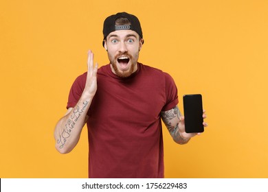 Surprised bearded tattooed man guy in casual t-shirt black cap isolated on yellow background. People lifestyle concept. Mock up copy space. Hold mobile phone with blank empty screen, spreading hands