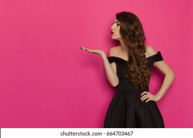 Surprised beautiful young woman in colorful vibrant striped mini dress is holding hands raised, presenting something, looking at camera and shouting. Studio shot on turquoise background.
