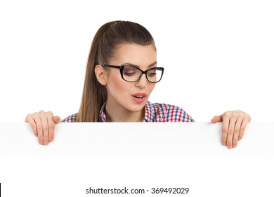 Surprised beautiful young woman in glasses and lumberjack shirt peeking behind white placard and looking down. Head and shoulders studio shot isolated on white.