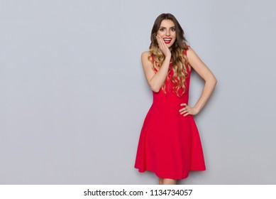 Surprised beautiful young woman in red dress is holding hand on chin and looking at camera. Three quarter length studio shot on gray background.
