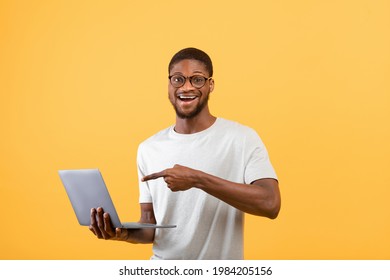 Surprised Black Man Pointing At Laptop Screen With Opening Mouth, Standing With Pc On Yellow Studio Background. Young Man Watching Unbelievable Breaking News And Shocking Web Content