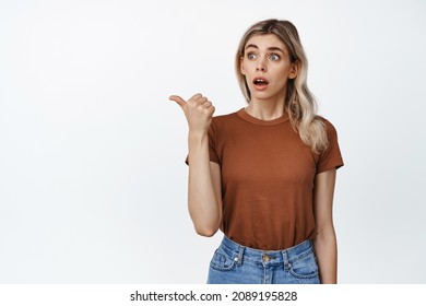 Surprised Blond Girl Gasp Impressed, Pointing Finger Left And Looking At Advertisement Amazed, Standing Against White Background