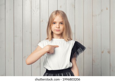 Surprised Blonde Teen Girl Points At Herself, Asks Who Me, Look Startled With Surprise News. Studio Shot, Light Wooden Background