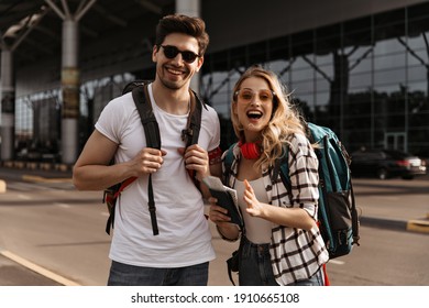 Surprised blonde woman and happy brunette man in sunglasses poses near airport. Cheerful travelers smiling and holding backpacks.