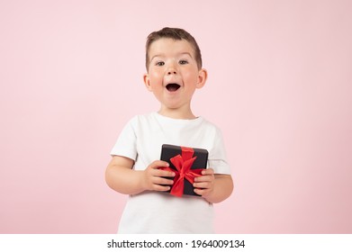 Surprised boy in white t-shirt holding black box gift in front, for girl or mother, Valentines day, mother's day. Pink studio background. High quality photo