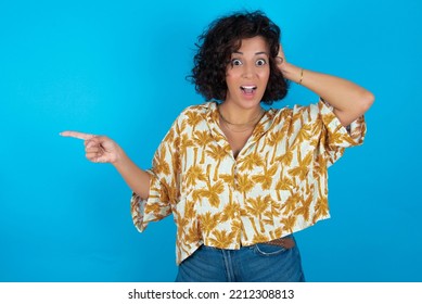 Surprised Brunette Arab Woman Wearing Hawaiian Shirt Over Blue Background Pointing At Empty Space Holding Hand On Head