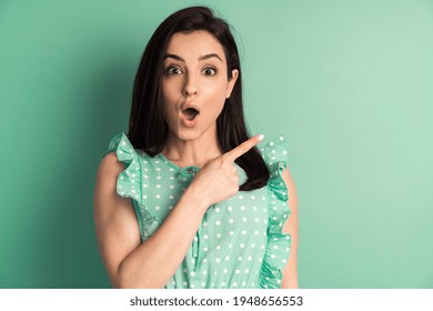 Surprised brunette and polka dot dress with index finger on copy space. Young woman looking at camera isolated on mint background.
