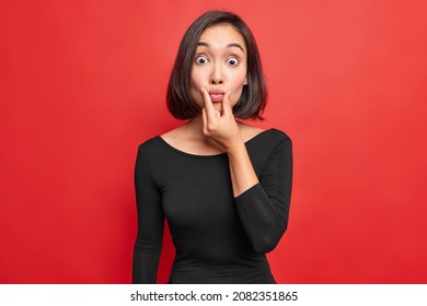 Surprised Brunette Young Woman Puckers Lips At Camera Looks Surprisingly Wears Black Dress Has Slim Figure Poses Against Vivid Red Background Looks Shocked Stands Indoor. Human Facial Expressions