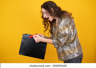 Surprised Caucasian Woman In Party Clothes Opening Gift Bag On The Yellow Background. Sales Concept. High Quality Photo