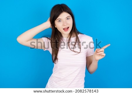 Surprised Caucasian woman wearing striped T-shirt over blue background pointing at empty space holding hand on head