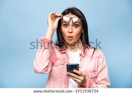 Surprised caucasian young pretty woman looks excitedly at the camera, takes off glasses, holds smartphone, received an unexpected message, saw interesting news, stands on isolated blue background