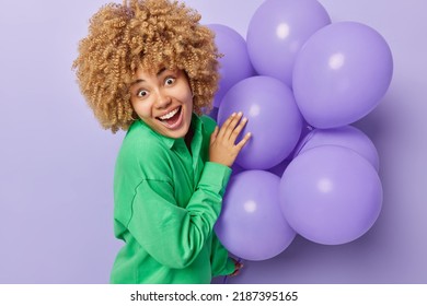 Surprised Cheerful European Woman With Curly Blonde Hair Wears Green Jumper Holds Bunch Of Inflated Balloons Celebrates University Leaving Prepares For Party Isolated Over Purple Background.