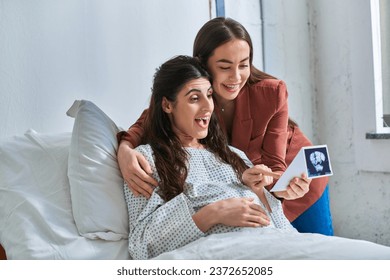 surprised cheerful lgbt couple looking at ultrasound of their baby with hands on belly, ivf concept