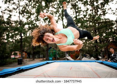 surprised cheerful woman with curly hair actively jumping on big trampoline. Green trees in the background. - Powered by Shutterstock