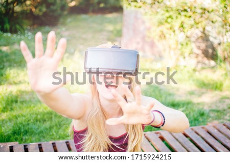 Surprised cheerful Woman using 3D  VR-virtual reality headset. Blonde thrilled Woman gesturing while using digital entertainment outdoors in green nature. Happy young female looking 3D digital movie  