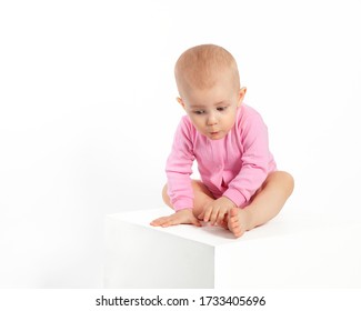 The surprised child. The baby sits sideways on a white cube and looks down in surprise. The little girl in pink pursed her lips. Beautiful children, cute European girl.