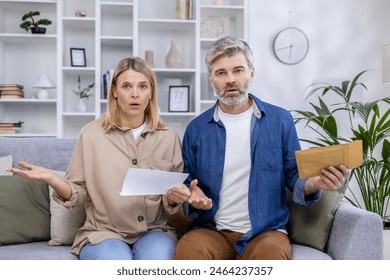 Surprised couple sitting on their couch, holding bills and documents in their living room, shocked by unexpected expenses or financial issues. - Powered by Shutterstock