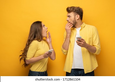 Surprised Couple With Smartphones Looking At Each Other On Yellow Background