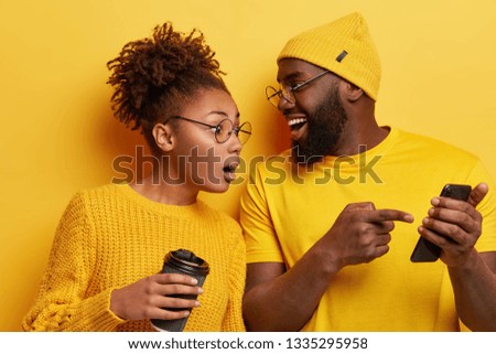 Surprised curious dark skinned woman looks at screen of boyfriends cellular, reads online post with interesting content, excited with smartphone or app features, isolated over yellow background.