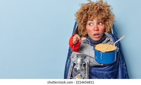 Surprised Curly Female Tourist Woman Hiker Holds Lantern And Pot Of Noodle Feels Cozy And Warm Wrapped In Sleeping Bag Feels Amazed Concentrated Aside Isolated Over Blue Background Blank Space