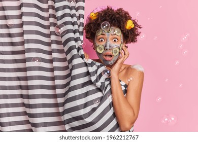Surprised curly haired woman takes shower in bathroom applies clay mask and cucumber slices for nourishing skin isolated over pink background hides naked body behind curtain. Skin care concept