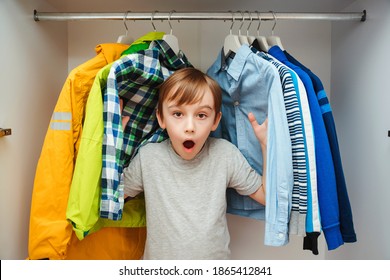 Surprised Cute Boy Searching For Clothing In A Closet. Preteen Boy Chooses Clothes In The Wardrobe Closet At Home. Kid Hiding Among Clothes In Wardrobe.