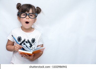 Surprised Cute Child In Eyeglasses,  Writing In Notebook Using Pencil, Keeping Mouth Wide Open. Four Years Old Kid, Isolated On White, Space For Advertising Text