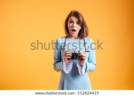 Surprised cute young woman standing with mouth opened and holding old vintage camera over yellow background