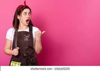 Surprised Dark Haired Woman Makes Gesture With Her Thumb. Gifted Active Baker Holds Kitchen Equipment In Right Hand, Opens Her Mouth And Big Eyes Widely, Wears Work Clothing. Cooking Concept.